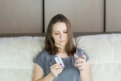 A woman is sat holding a blister pack of painkiller tablets & a glass of water, she is suffering from painkiller dependency
