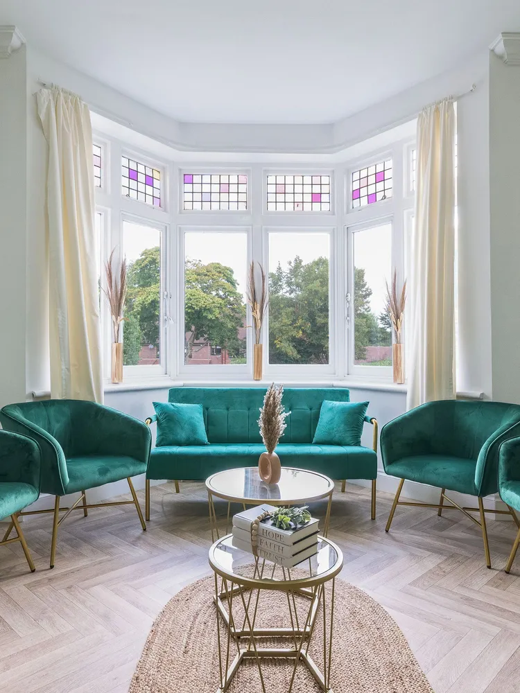 Bright and elegant sitting room with green velvet chairs, gold-legged tables, and a large bay window featuring stained glass panels and cream curtains.