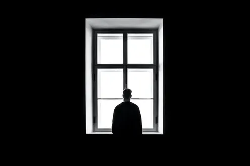 A lonely mans silhouette stands against a large framed window looking out to the distance