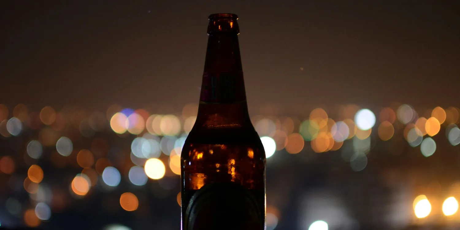 A beer bottle silhouetted against blurred city lights at night, symbolising alcohol misuse, loneliness and the cycle of drinking and depression.