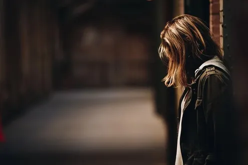 A side profile of a person who stands against a wall, they are looking down with their long hair covering their face