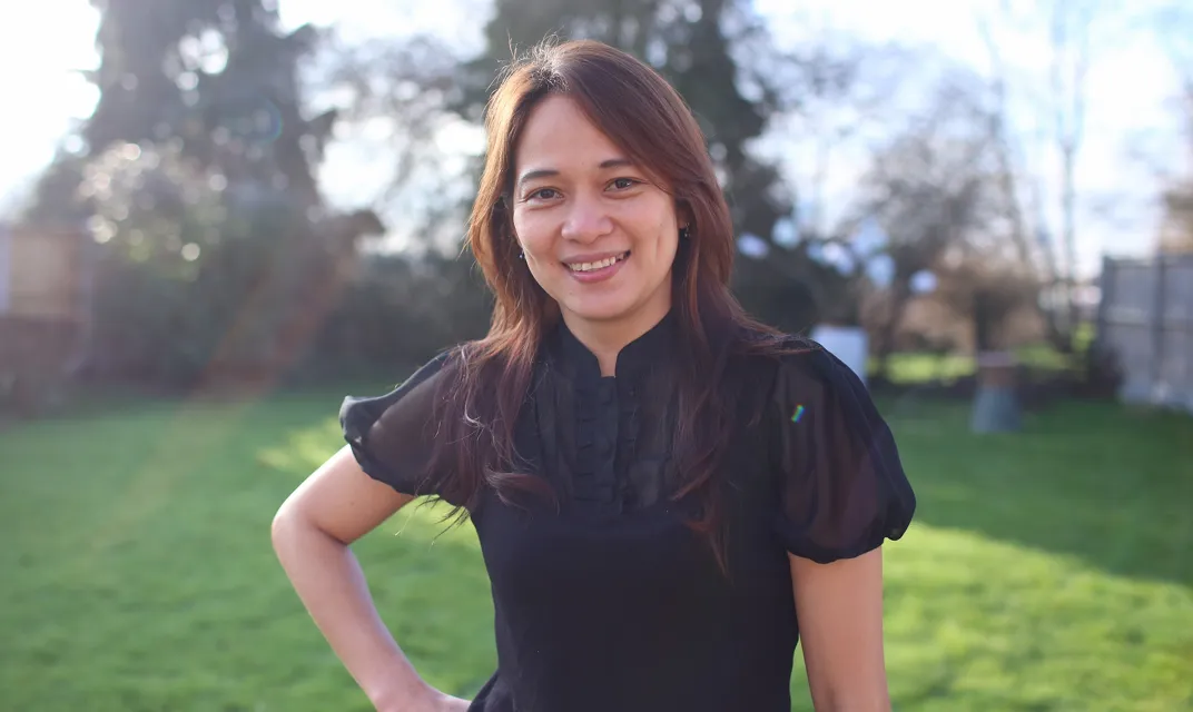 Jenny Esmas wearing a black short-sleeved blouse, stands smiling outdoors on a sunny day with green grass and trees in the background.