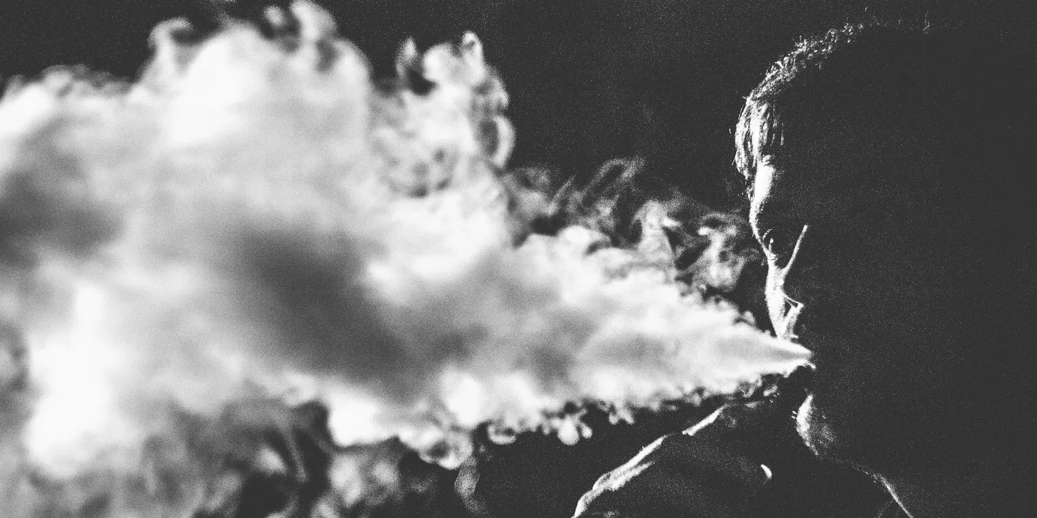 black and white image of a man blowing thick plumes of vape smoke.