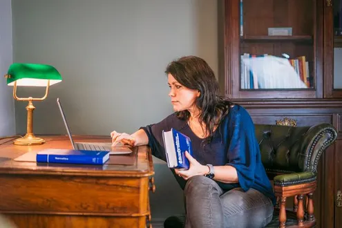 Rehabs UK BACP Therapist Jakana Rita sits in an armchair at a desk in a stylish office, consulting books and a laptop.