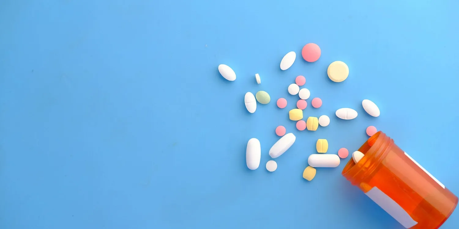 ssorted tablets and capsules spilled from an orange prescription pill bottle onto a blue background.