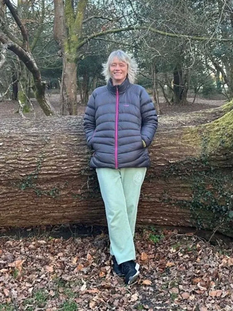 Woman in a dark jacket and light green trousers leaning against a large fallen tree in a woodland area, surrounded by leaf-covered ground and tall trees