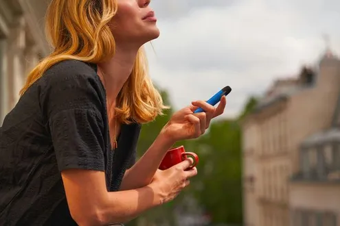 Woman holding a blue vape device and coffee cup while sitting outdoors on a balcony in an urban setting.