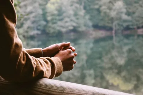 Clasped hands rest on a wooden railing with water and trees in the background representing obsessive thinking about drugs
