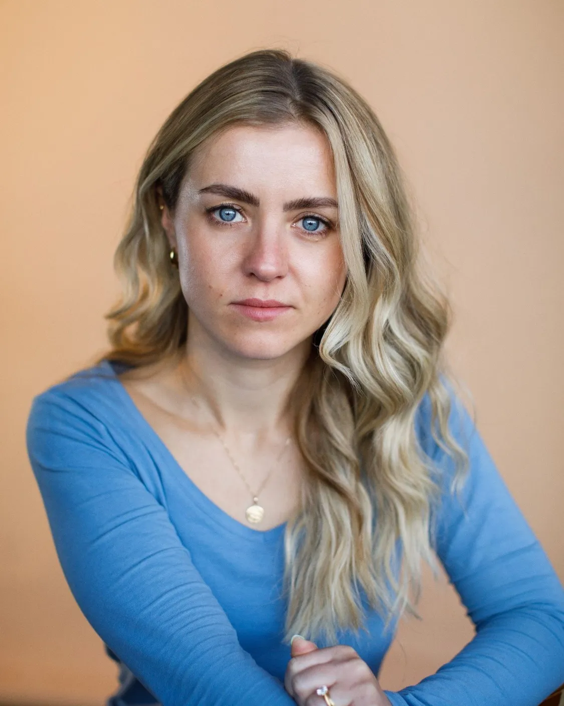 Confident young woman with long blonde hair and blue eyes wearing a light blue top, sitting indoors against a neutral background, representing professionalism and approachability.