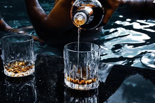 Two crystal tumblers sit on a shiny bar table, one of the tumblers has alcohol in it and the other one is being poured into