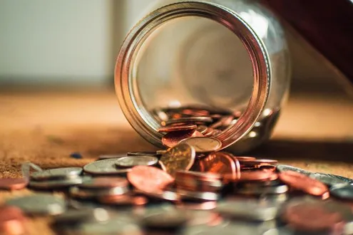 A glass jar has fallen on the floor, it contains coins that are spilling out of it