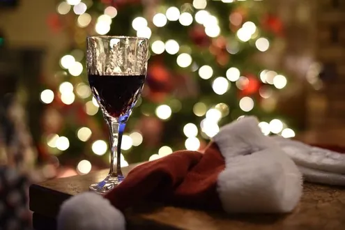 A glass of red wine sits on a table next to a Santa's hat, in the background there is a Christmas tree with lights