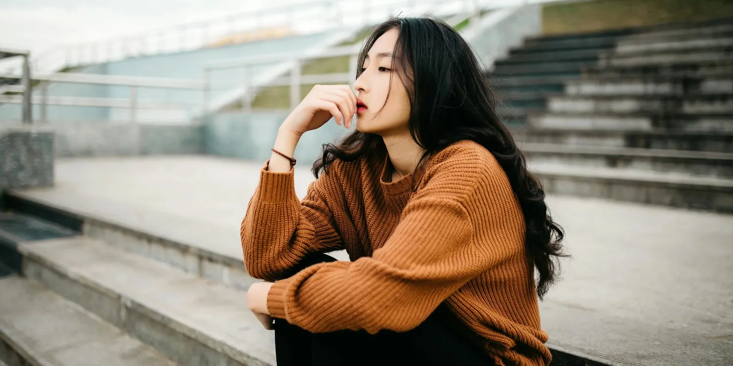 Young woman in a brown knitted jumper sitting on outdoor concrete steps with a thoughtful expression, creating a calm and reflective mood.