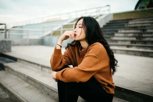 Young woman in a brown knitted jumper sitting on outdoor concrete steps with a thoughtful expression, creating a calm and reflective mood.