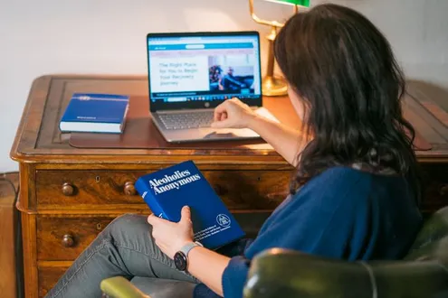 Jakana Rita BACP Therapist at Rehabs UK is sat at her desk with her laptop holding the Alcoholics Anonymous book in her hand