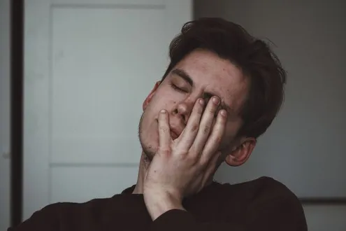 A man is sat with his hand against his face suffering from a migraine which is linked to alcohol use