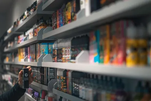 A person is reaching for a vape off a shelf in shop highlighting the increase of vaping in use of NOS in the UK