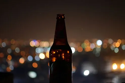 A beer bottle silhouetted against blurred city lights at night, symbolising alcohol misuse, loneliness and the cycle of drinking and depression.