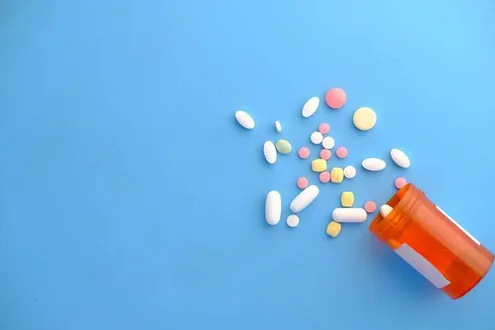 ssorted tablets and capsules spilled from an orange prescription pill bottle onto a blue background.