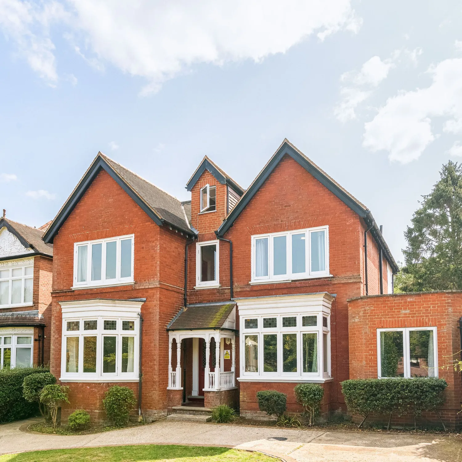 Front exterior view of a large red-brick detached Victorian-style house with white-framed windows, a gabled roof, and a small front garden on a sunny day.