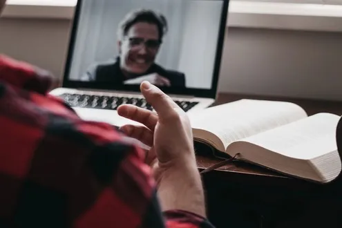 A man sits at a table facing his laptop engaging in online therapy with his counsellor, a book is opened to his side