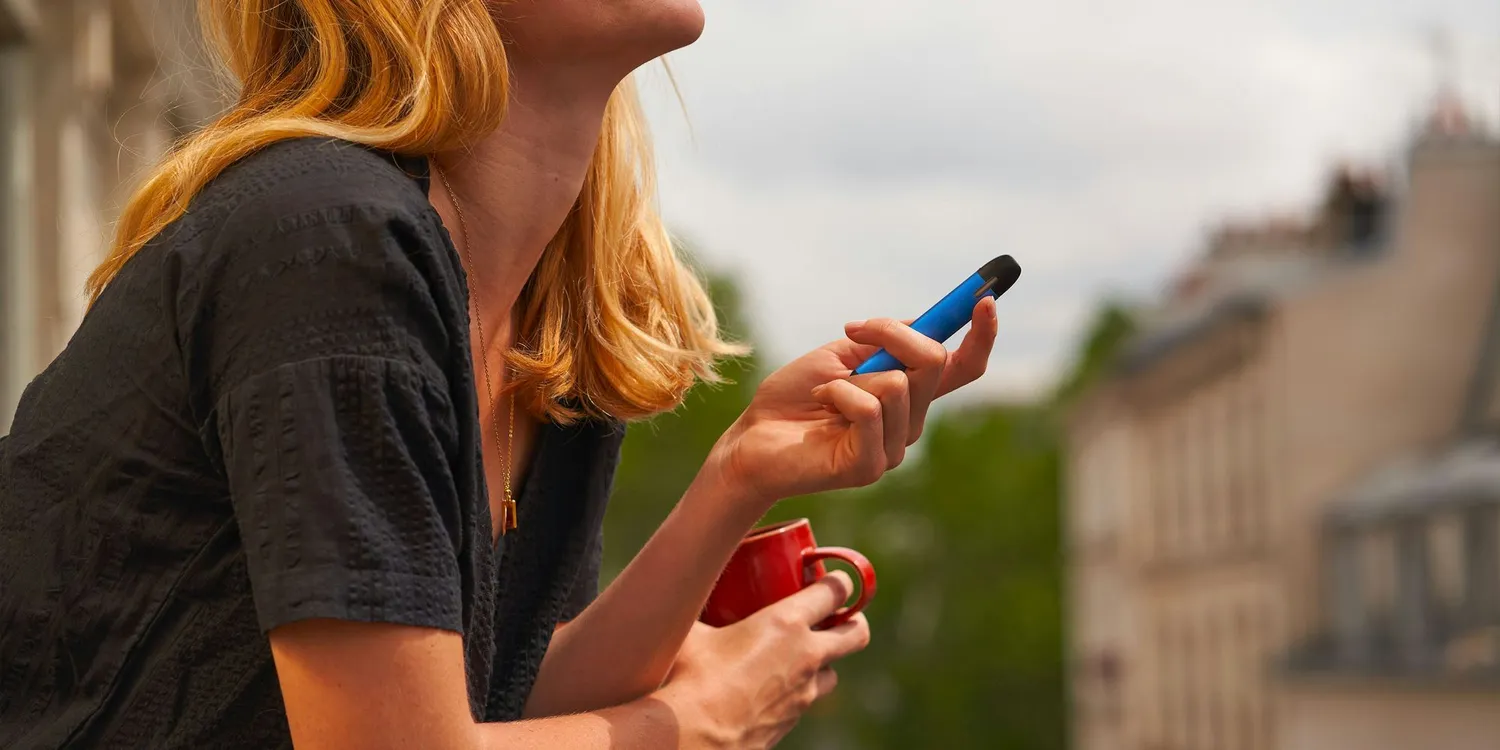 Woman holding a blue vape device and coffee cup while sitting outdoors on a balcony in an urban setting.