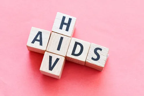 Separate wooden blocks with letters on them are sat on a pink table it spells HIV vertically and AIDS horizontally