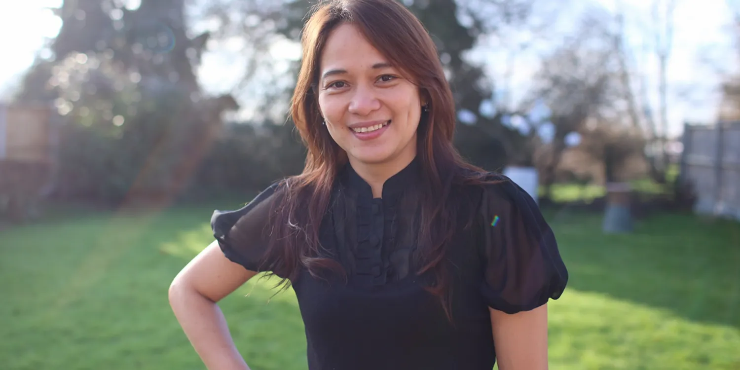 A woman with long brown hair, wearing a black short-sleeved blouse, stands smiling outdoors on a sunny day with green grass and trees in the background.