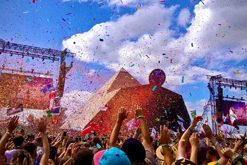 A crowd at Glastonbury Music Festival Pyramid stage raise their arms in the air whilst coloured confetti showers them