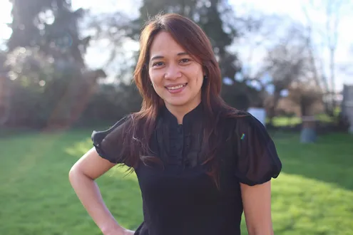 A woman with long brown hair, wearing a black short-sleeved blouse, stands smiling outdoors on a sunny day with green grass and trees in the background.